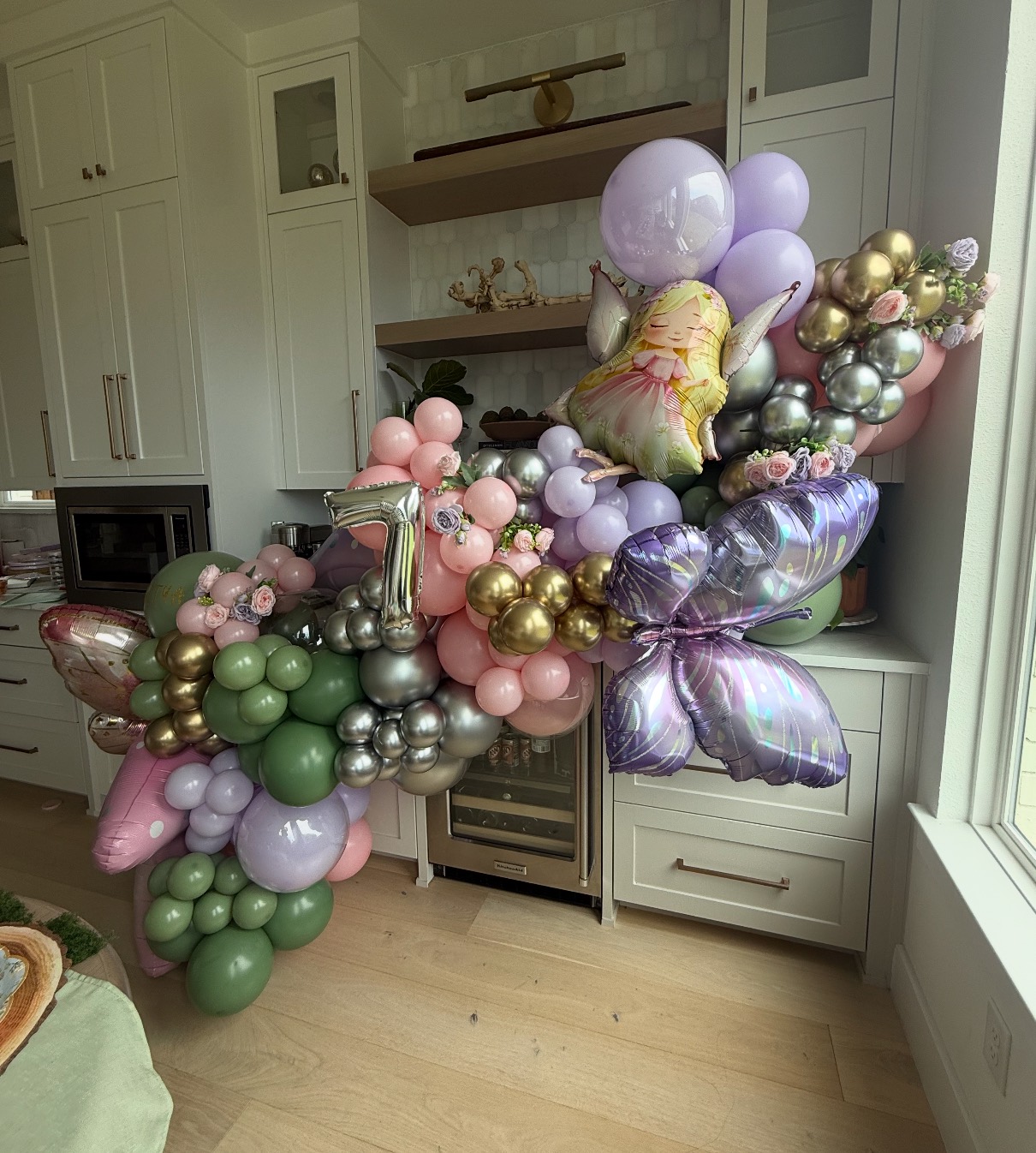 A sage, blush and lilac garland threaded with chrome and gold around a kitchen bar, hung with butterfly foils, a fairy and a silver number seven