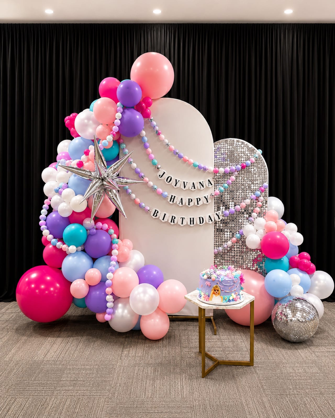 Pink, violet and sky-blue balloons banked around a white arch and a silver sequin wall, under a chrome starburst